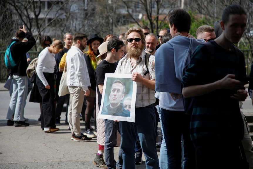 A long line of people standing outdoors, including one holding a picture with a man's face on it