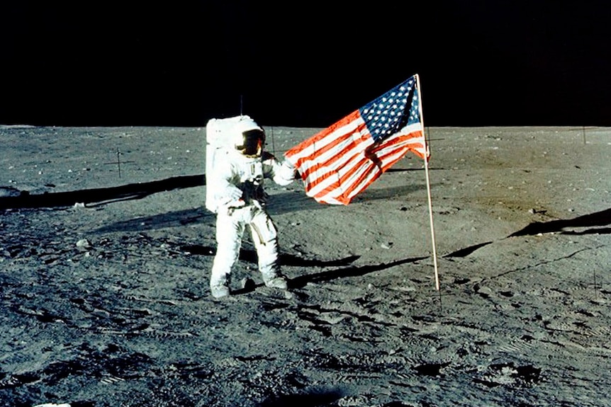 An astronaut stands on the surface of the moon in a white spacesuit holding the corner of an American flag.