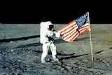 An astronaut stands on the surface of the moon in a white spacesuit holding the corner of an American flag