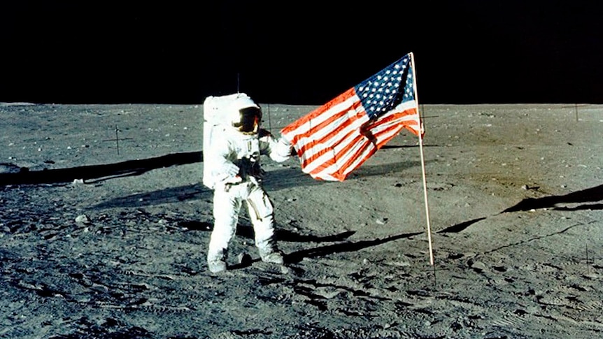 An astronaut stands on the surface of the moon in a white spacesuit holding the corner of an American flag