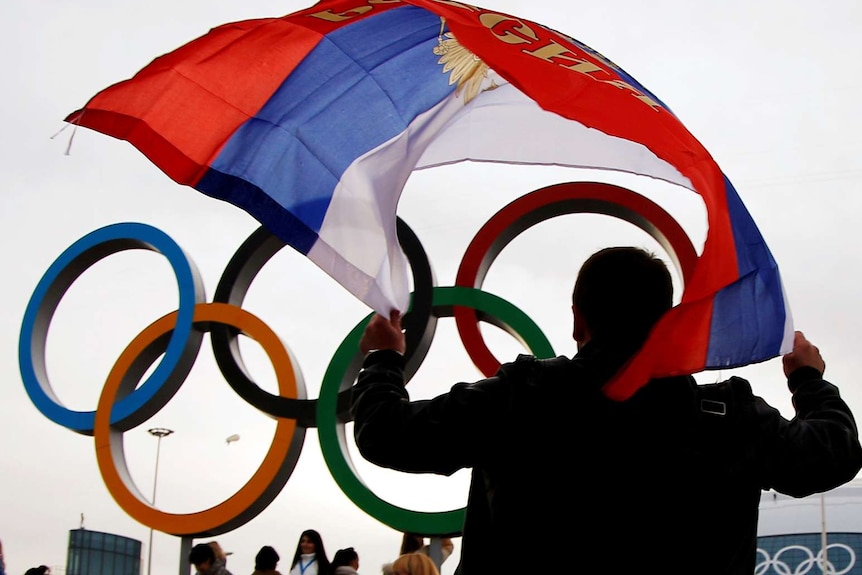 A person, with back to camera, holds a Russia flag above their head. The Olympic rings are behind him.