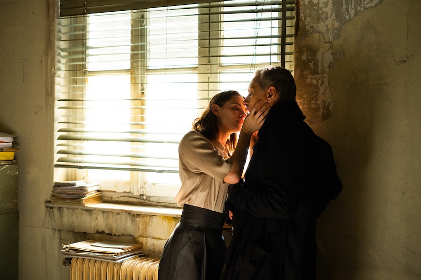 Brunette white woman in white blouse lifts face to white man in black coat as if about to kiss him.