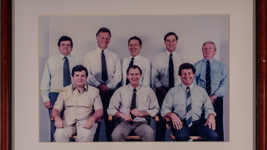 Eight men wearing shirts and ties smile. Four men at the back are standing, three sitting in chairs at the front.