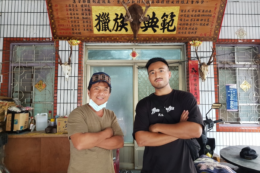 两名男子双手抱胸站在一块匾额下，匾额上用繁体中文书写“猎族典范”