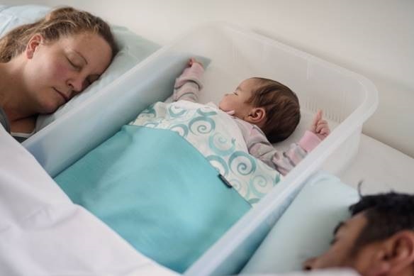 A baby sleeps inside a clear plastic tub with a man and woman on either side. they're all in the bed.