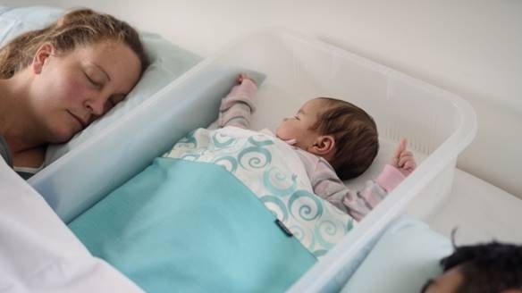 A baby sleeps inside a clear plastic tub with a man and woman on either side. they're all in the bed.