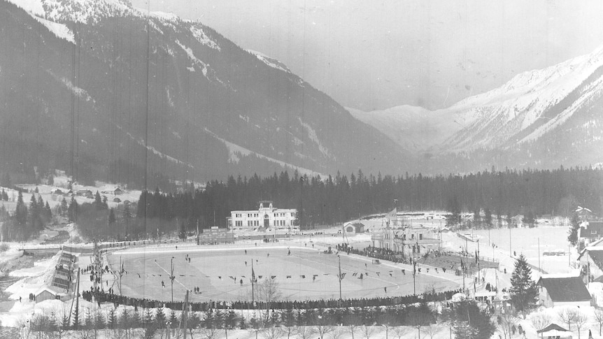 A black and white photo of a winter stadium with mountains in the background. 