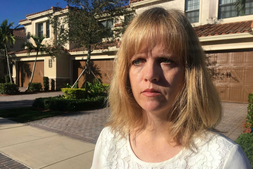 A woman stands in front of a two-storey home and looks sternly off camera