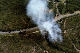 Smoke rises from lush green bushland near a bitumen road, as seen from above.
