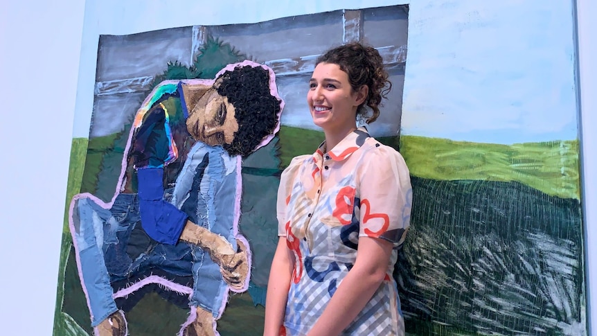 A woman smiles broadly as she stands in front of a large piece of textile art.