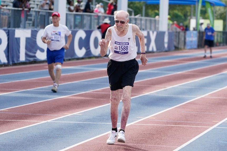 A 95-year-old man in a white singlet and black shorts powers down a running track in jogging shoes.