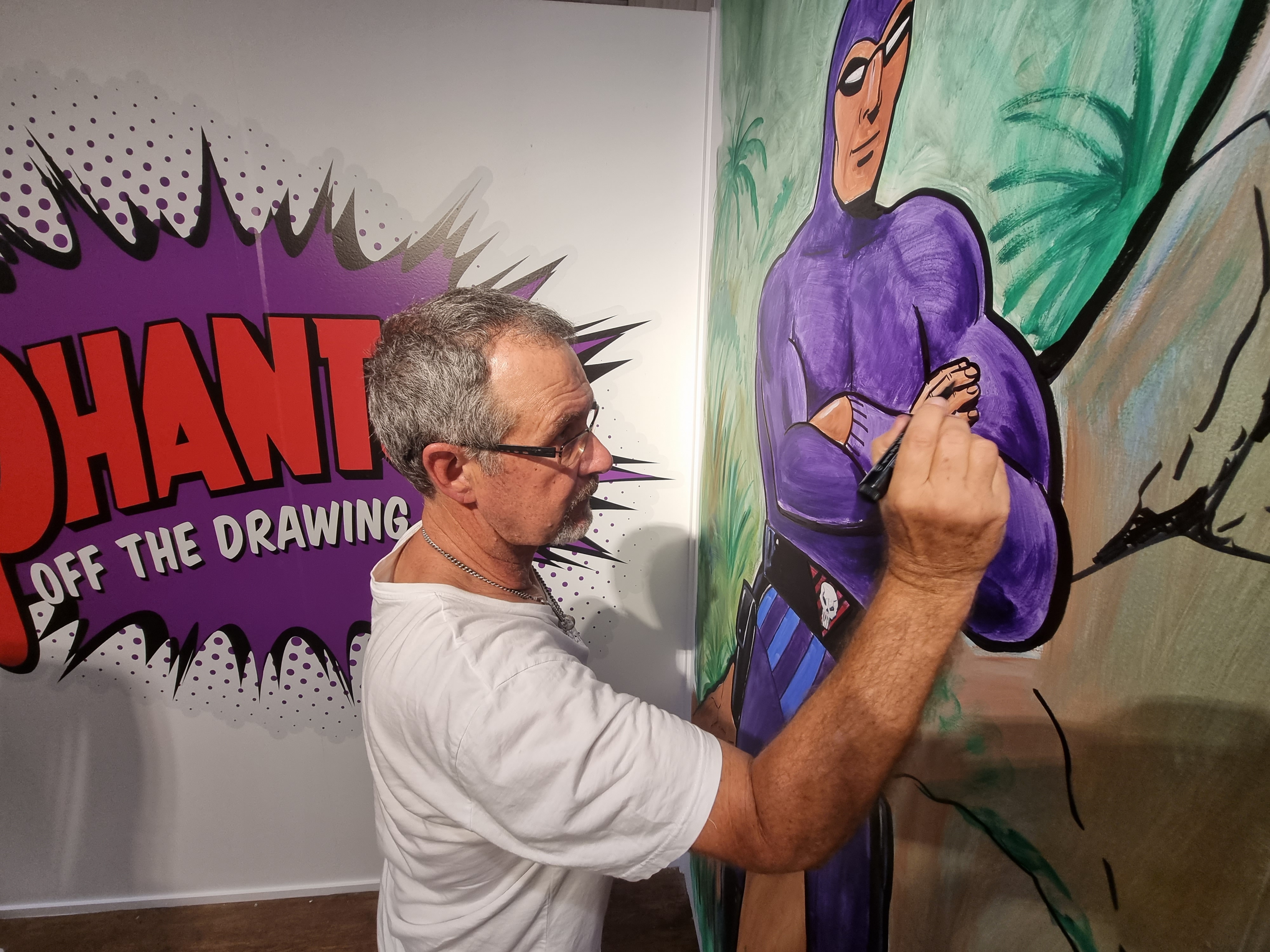 A man painting a mural on a large wall. The mural is a giant purple superhero, The Phantom