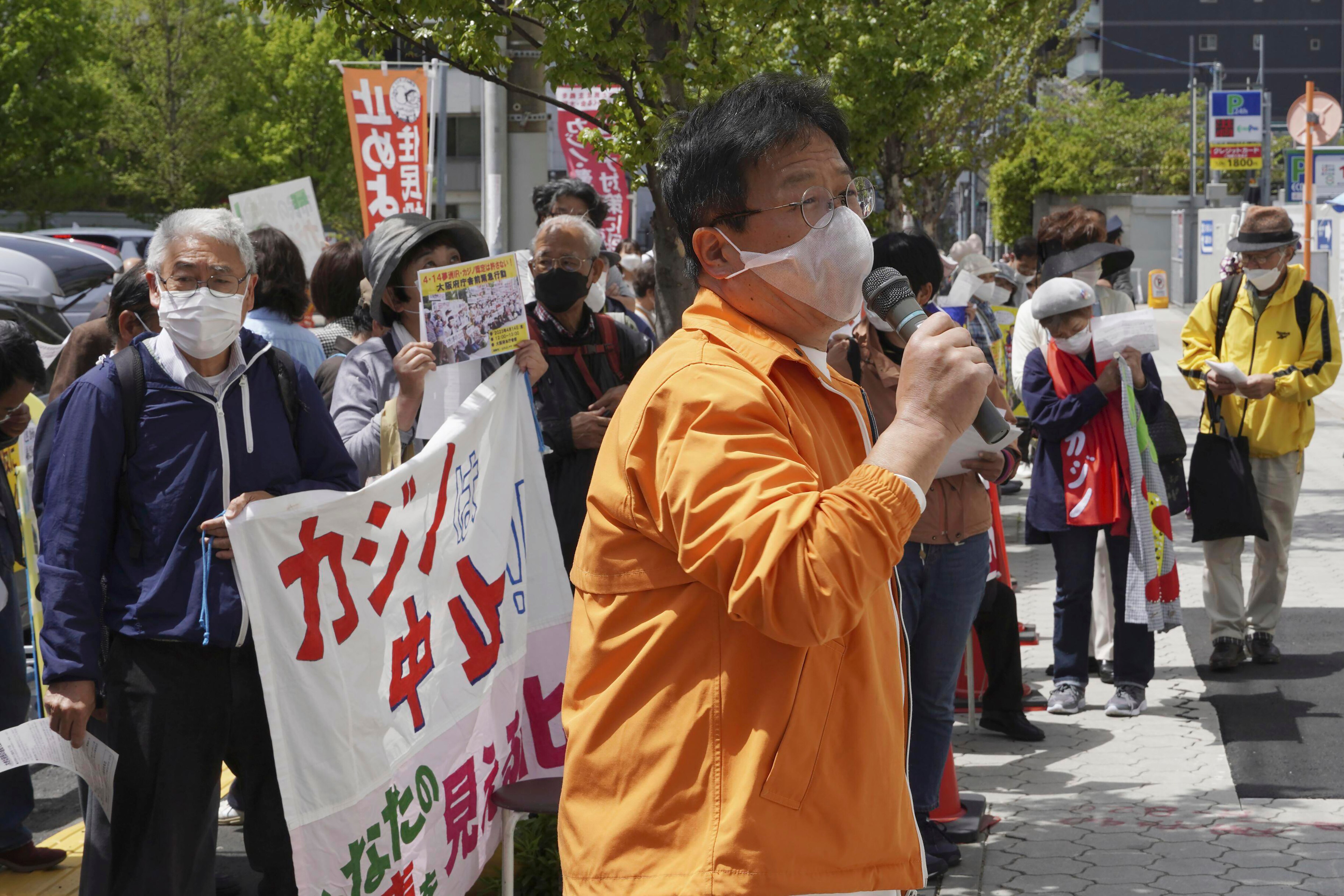 A man in an orange jacket wears a mask and speaks into a microphone as others hold signs.