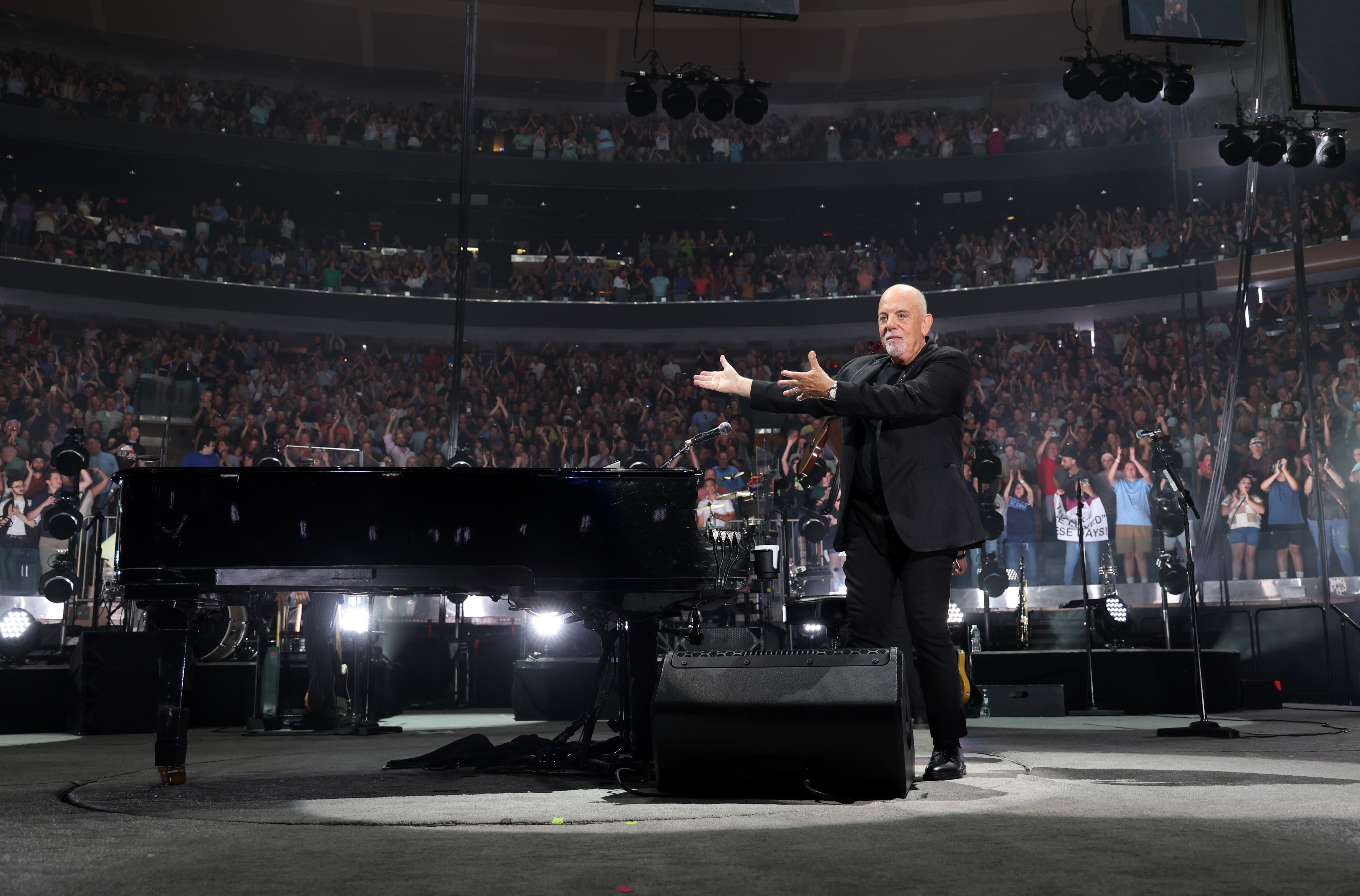 Billy Joel on stage with his piano, an enormous crowd seated in stands behind him.