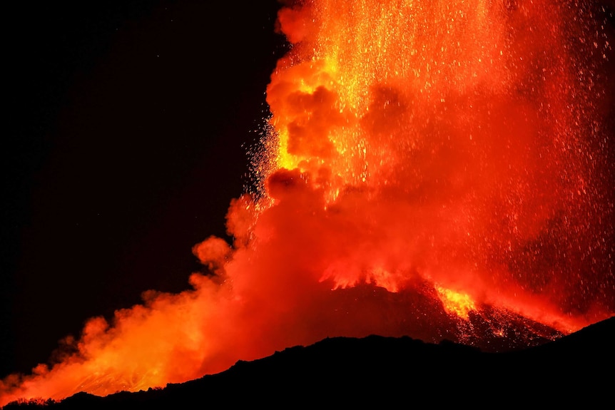 A wide shot of a mountain erupting at night with red hot lava shooting up.