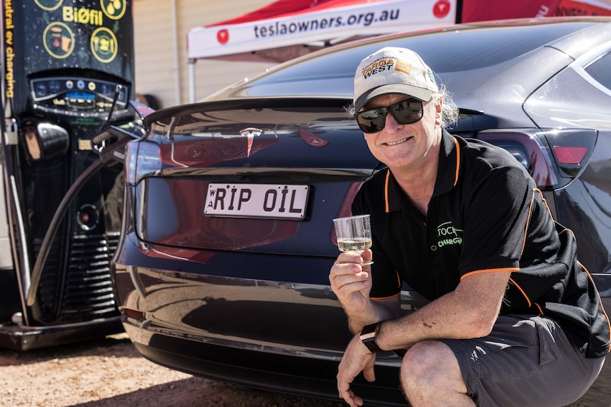 A man holding a plastic cup of champagne kneeling down next to his car.  