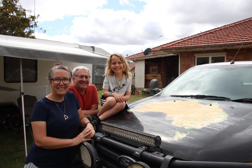 A woman, man, and child lean on the front of their ute in front of a brick house.