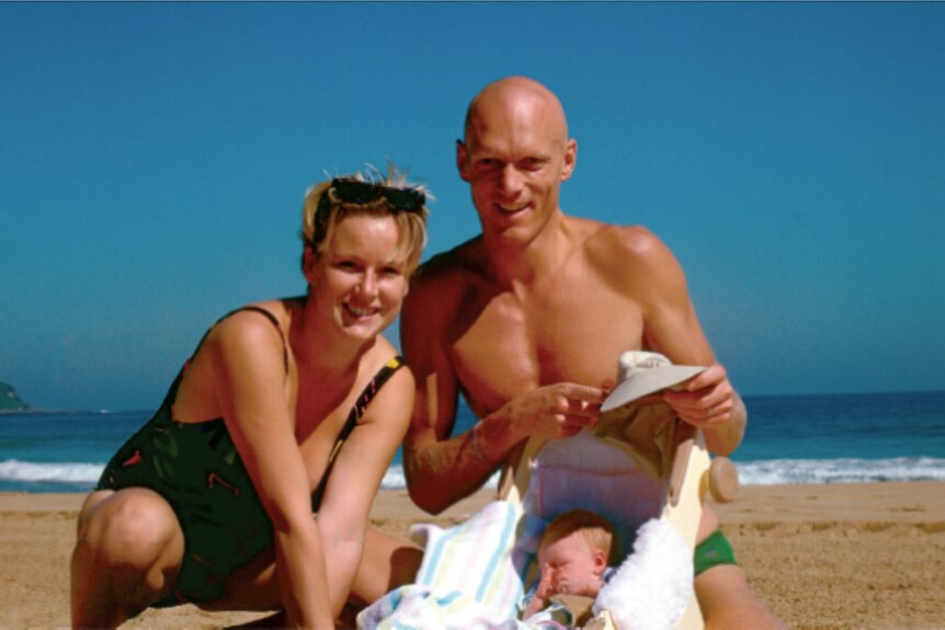 Young bald man and woman sit with their baby, in a carrier, on the beach