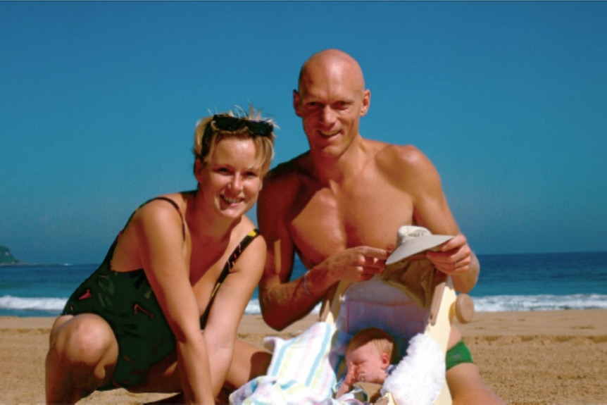 Young bald man and woman sit with their baby, in a carrier, on the beach