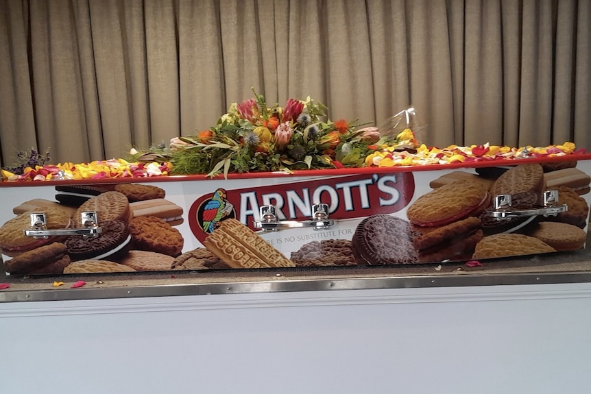 A coffin print with an Arnott's biscuit packet with a big bunch of flowers and petals sitting on top.