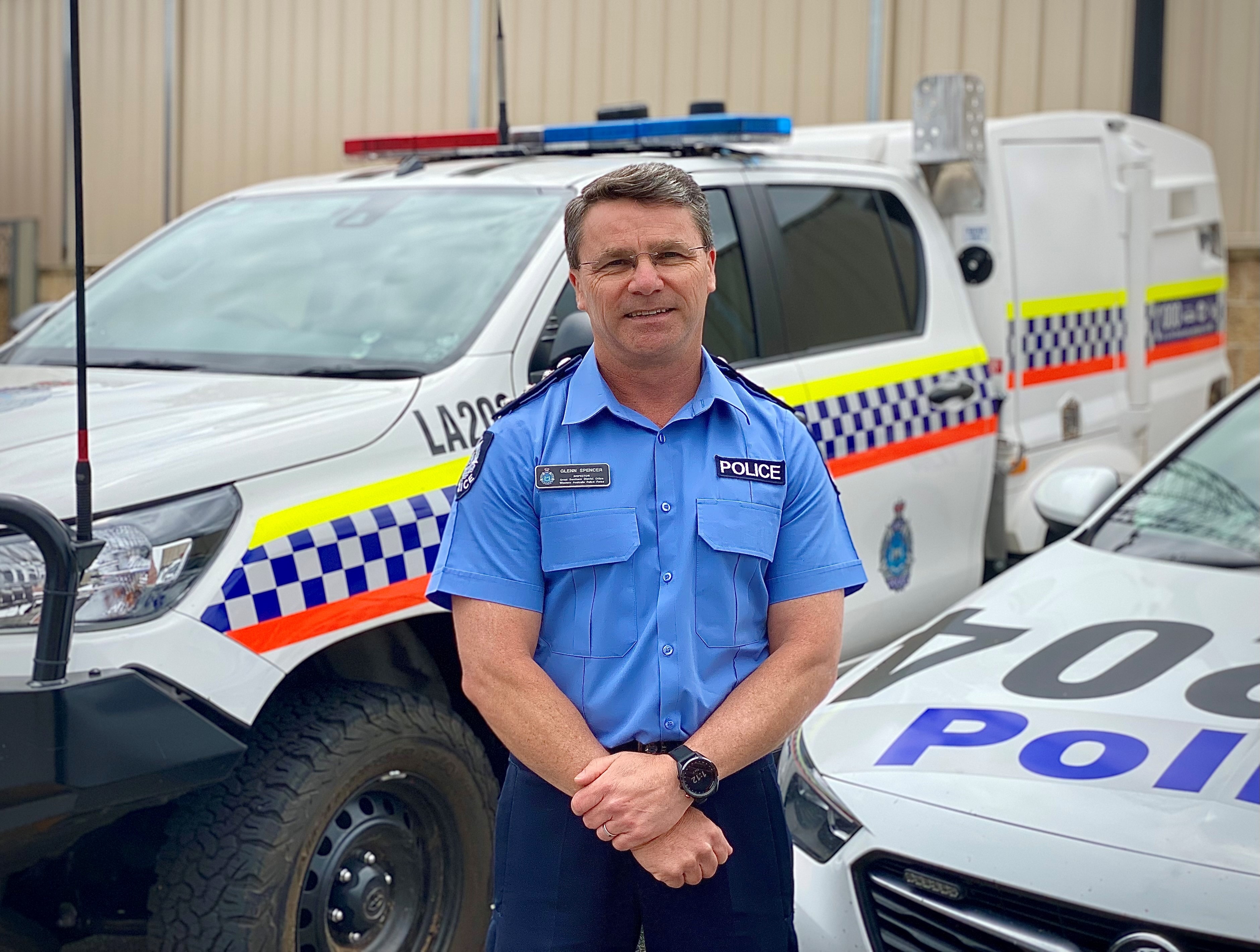 Inspector Glen Spencer stands with hands held in front.  In the background are two police vehicles. 