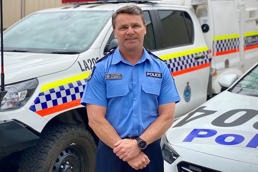 Inspector Glen Spencer stands with hands held in front.  In the background are two police vehicles. 