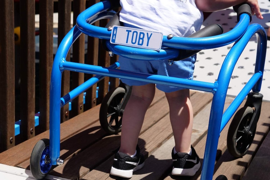 A little boy uses his blue walking frame on a playground.