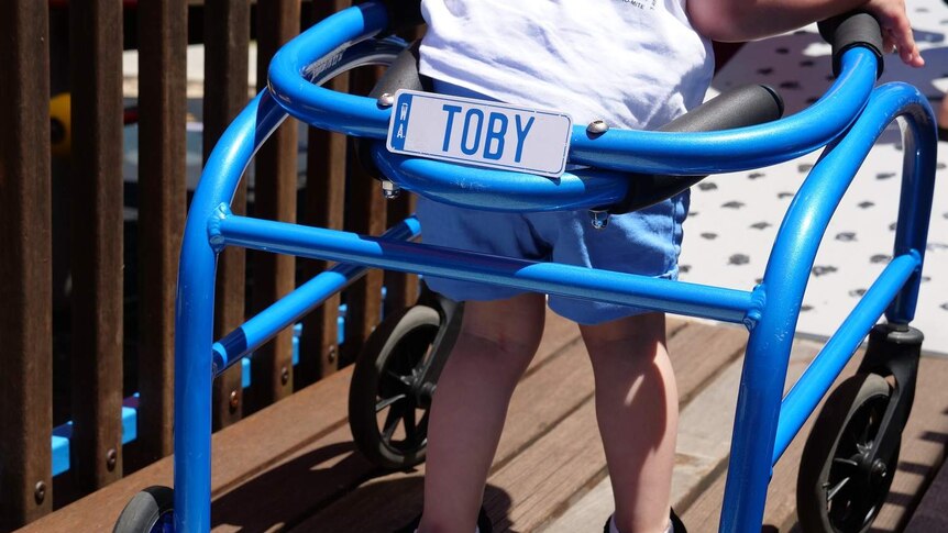 A little boy uses his blue walking frame on a playground.