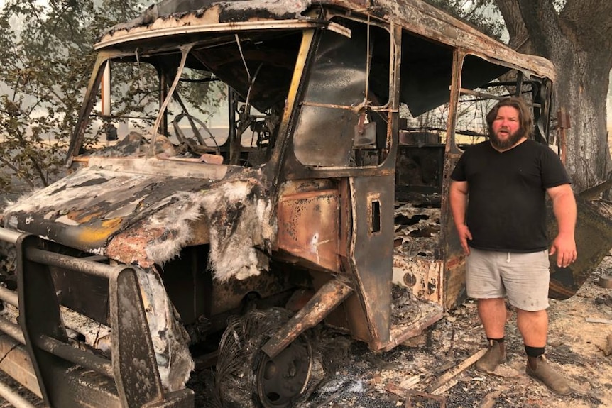 A young, bearded, long-haired man in a black shirt and shorts stands next to a burnt-out bus.