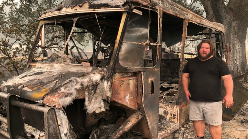 A young, bearded, long-haired man in a black shirt and shorts stands next to a burnt-out bus.