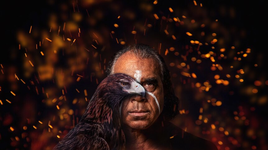 An Indigenous man with white ochre marking has half his face covered by a Wedgetail Eagle