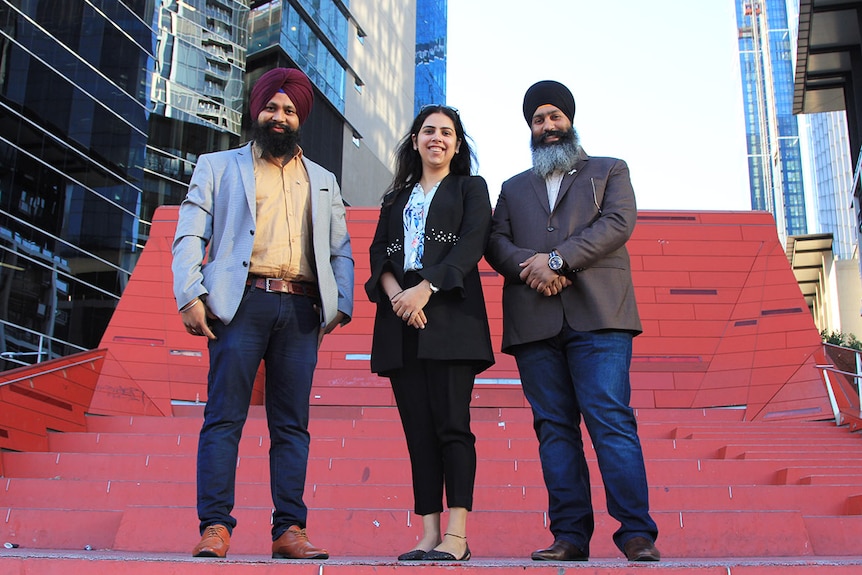 Sabhi Singh, Chaman Preet and Amar Singh stand in front of red stairs beside the highrise buildings at Melbourne's Southbank.