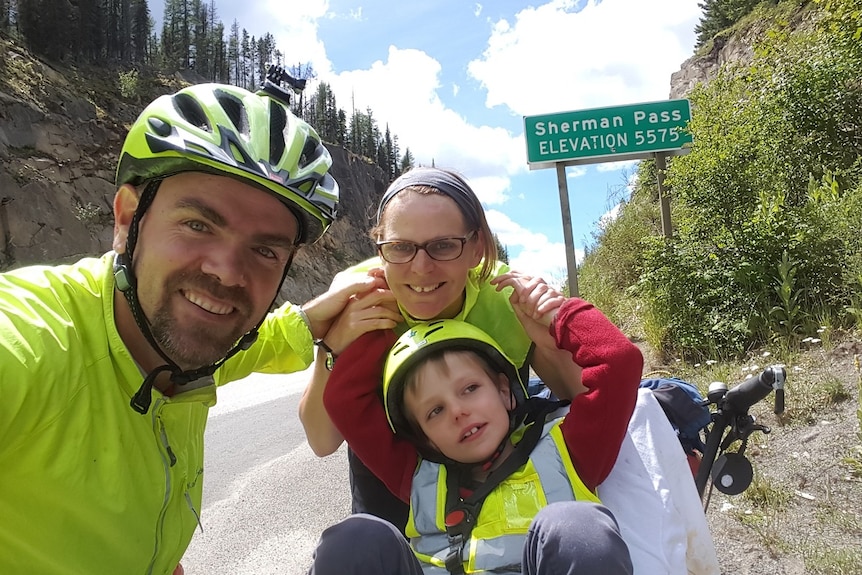Fiona Churchman, Travis Saunders and their son Patch in front of a road sign saying Sherman Pass