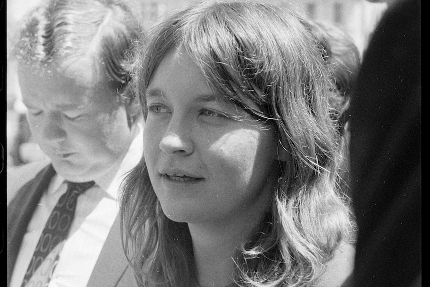 A young woman with long brown hair surrounded by reporters and being interviewed.