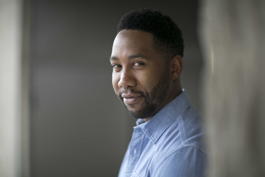 The side profile of a man, turned towards the camera, smiling and wearing a light blue shirt.