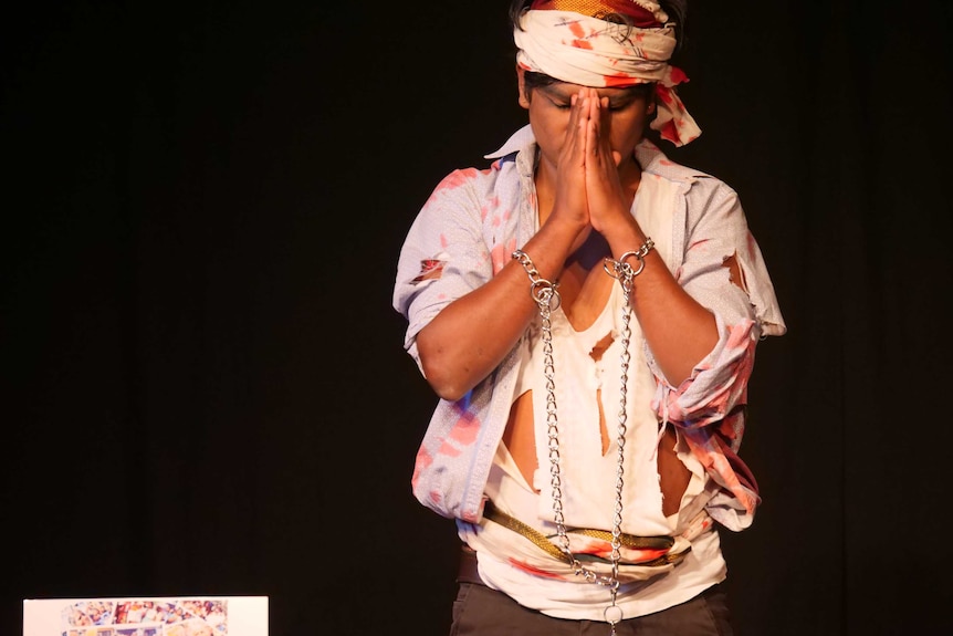 A man performing onstage in front of a black curtain wearing a ripped shirt and chains around his wrists bows head in prayer.