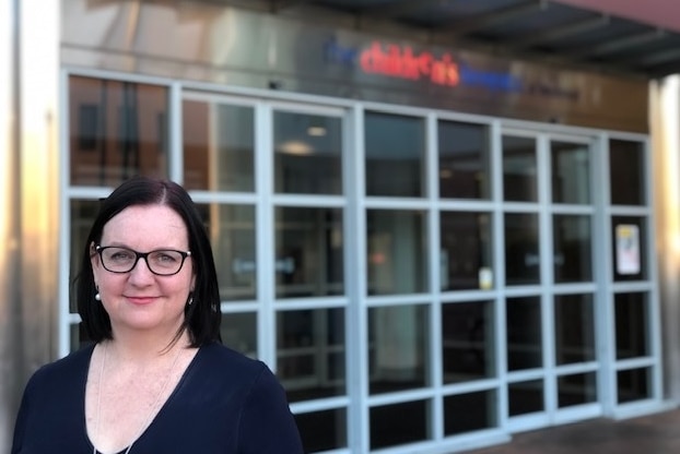 A woman wearing glasses stands in front of a sign and a set of glass doors.