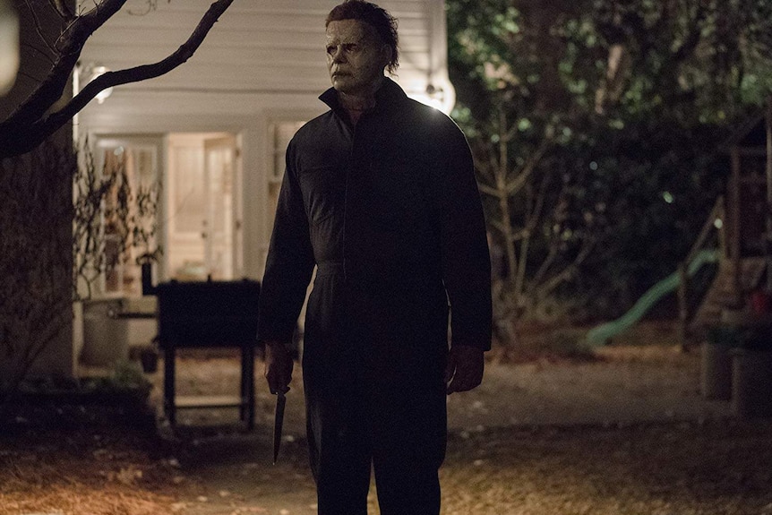 Michael Myers stands in a yard holding a knife wearing a mask