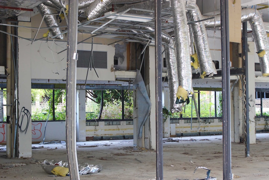 Silver ducting hangs from the ceiling in an abandoned building. There is graffiti on the walls and all the windows are smashed.