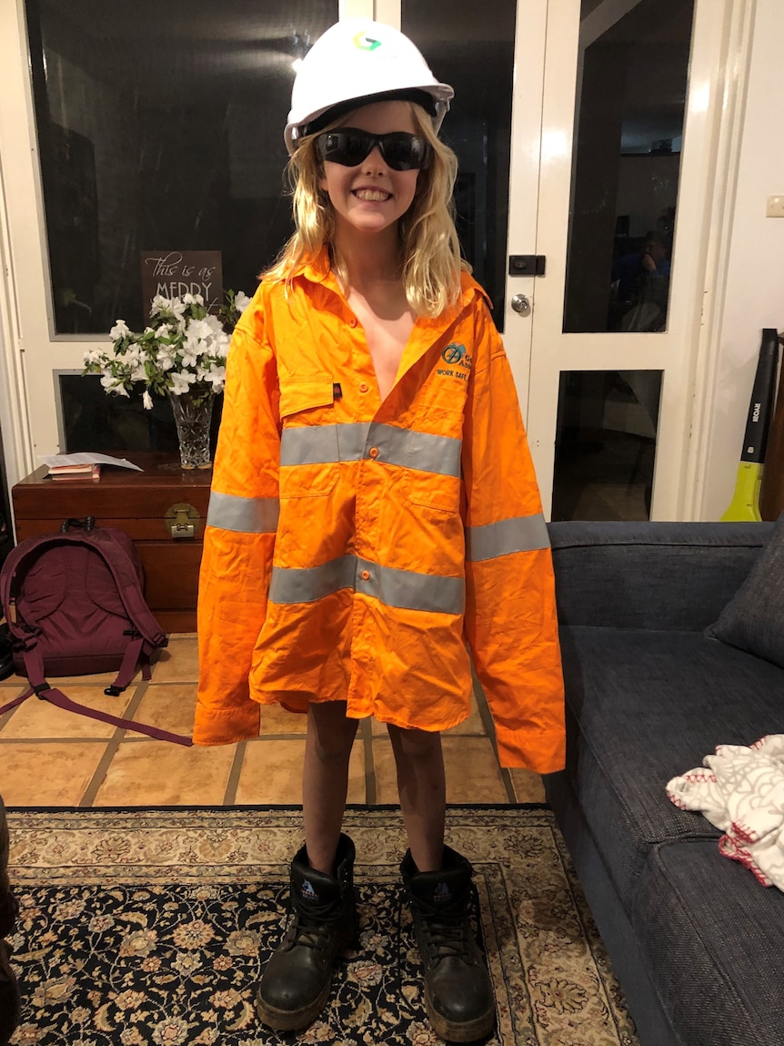 A young girl wears a high visibility mining jacket, hard hat and sunglasses