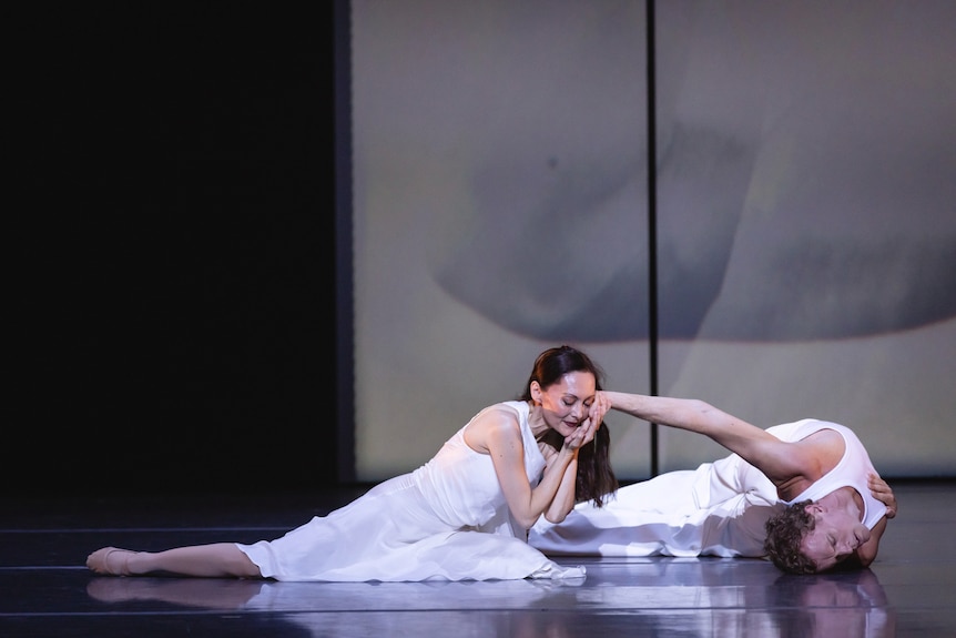 A male and female dancer on the floor of a stage. The man is stretched out and the woman holds his hand to her face.