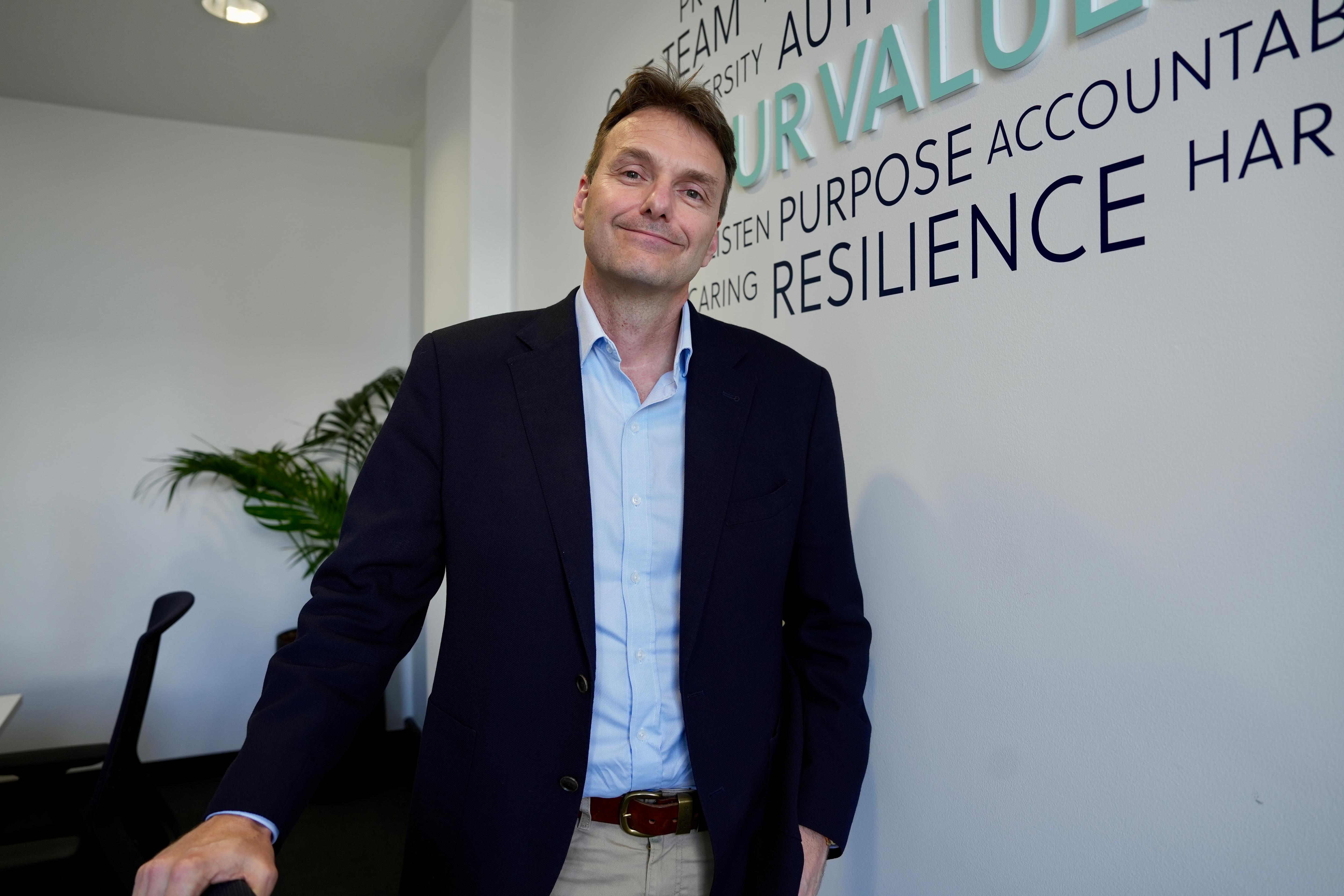 Man wearing blue shirt and dark blazer standing in front of a wall with themed words behind him
