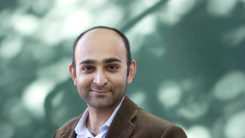 Colour photograph of author Mohsin Hamid standing in front outdoors with dappled light on a wall.