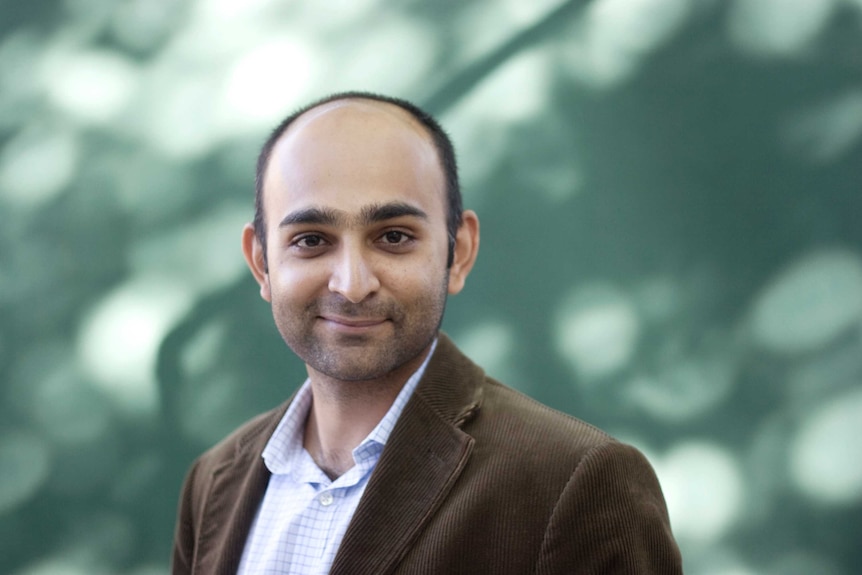 Colour photograph of author Mohsin Hamid standing in front outdoors with dappled light on a wall.