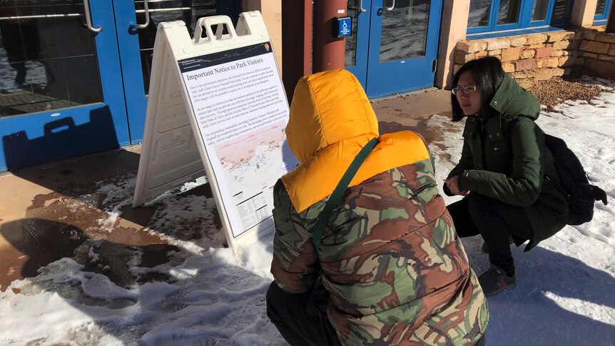 Two women kneel in the snow to read a notice posted on a signboard.