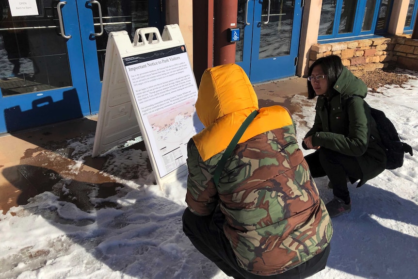 Two women kneel in the snow to read a notice posted on a signboard.