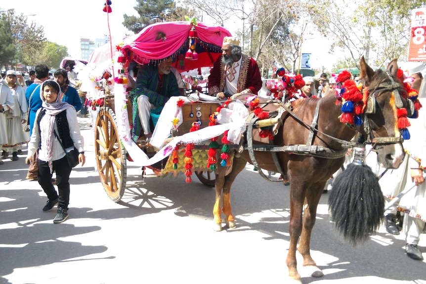 Dark haired woman walks alongside a horse-drawn carriage, surrounded by men in turbans 