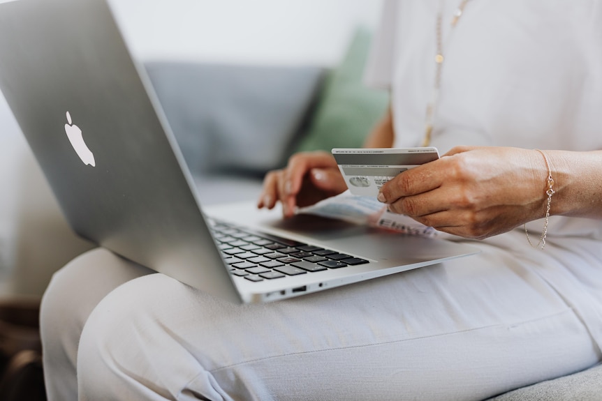 A brown woman wearing white jeans and a white top sits down with a laptop in one hand and a bank card in the other.