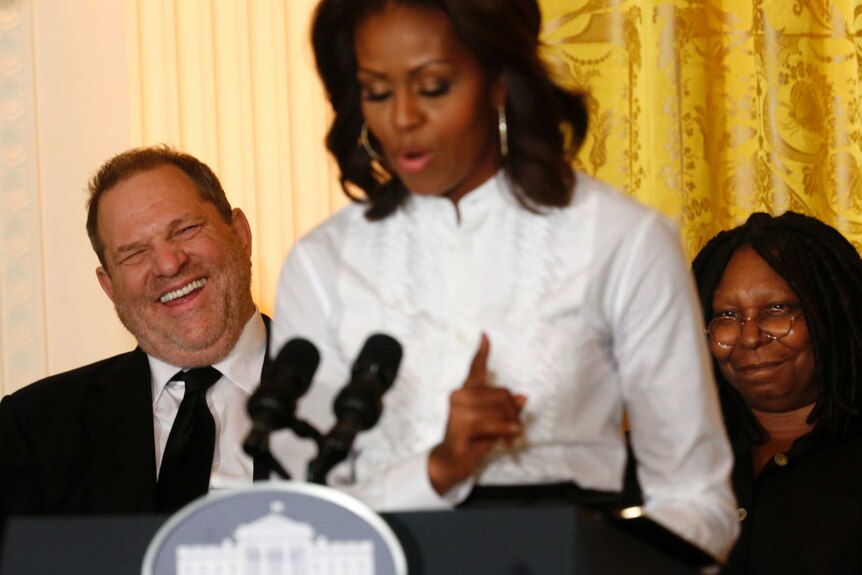 In-focus Harvey Weinstein laughs while sitting behind an out-of-focus Michelle Obama.