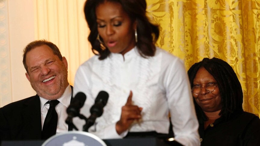 In-focus Harvey Weinstein laughs while sitting behind an out-of-focus Michelle Obama.